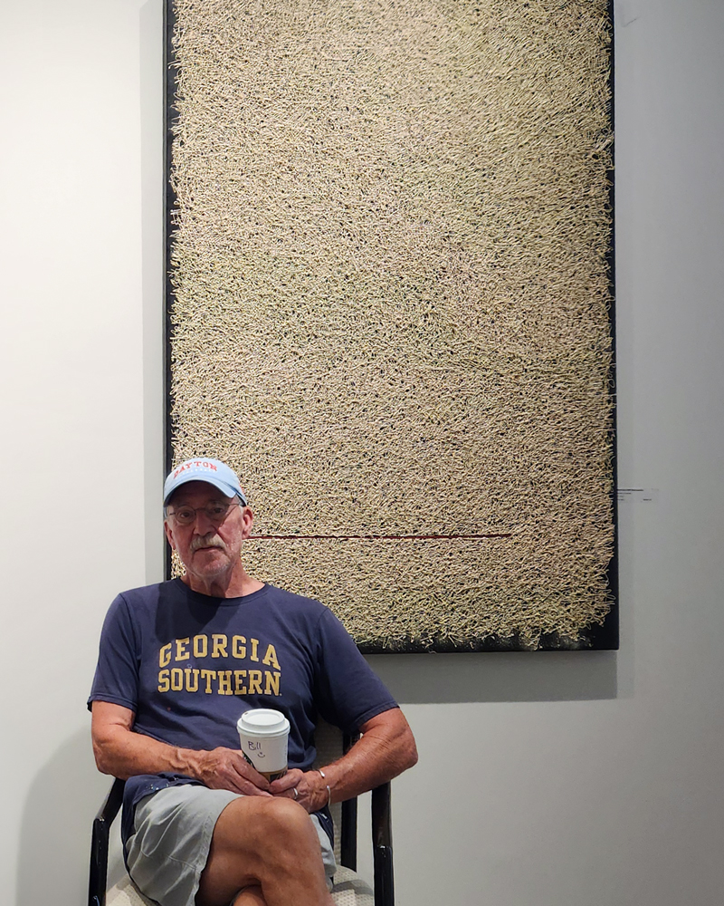 "Down Here With Everyone Else" with w.e. pugh sitting in front of the painting